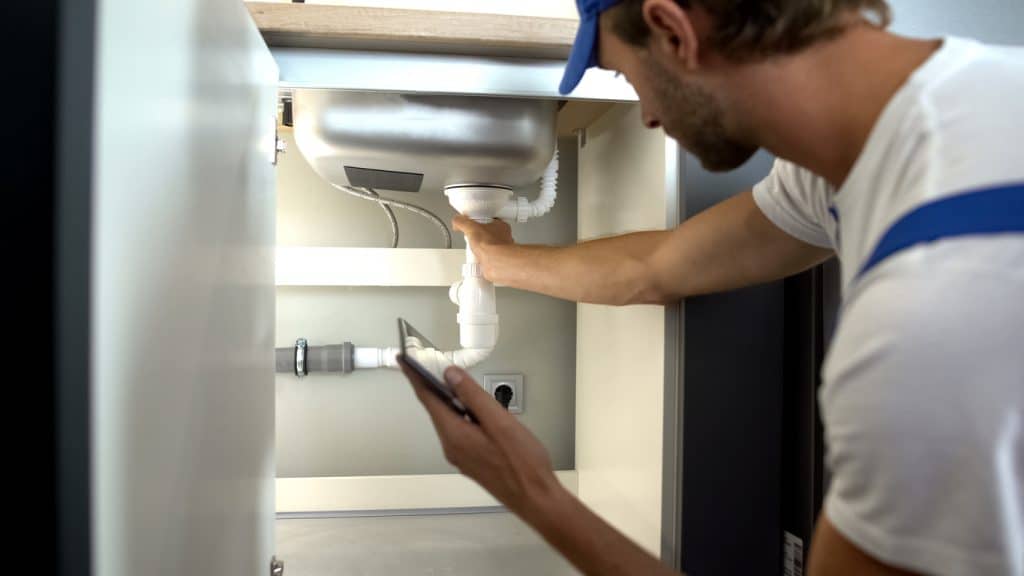 Professional plumber checking water pipes in kitchen, typing report in tablet