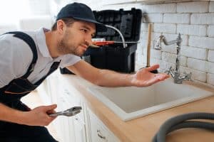 Young handyman check tap for leaking. He hold hand under it. Man stand in kitchen at sink. Toolbox and hose on desk